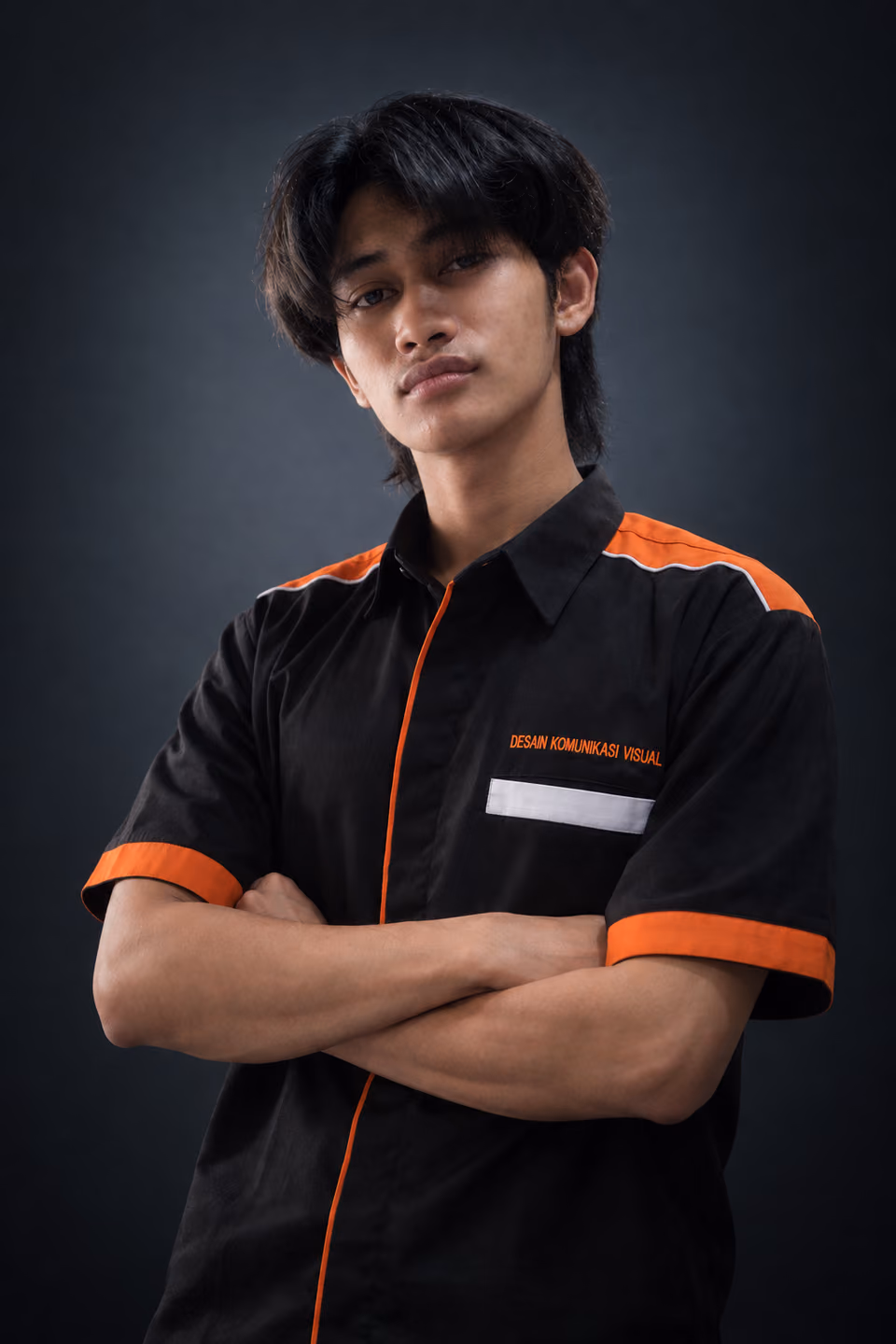 Ashabi Hastra mengenakan seragam DKV hitam beraksen oranye, bersedekap dengan pencahayaan rim studio dramatis pada latar bergradasi gelap.